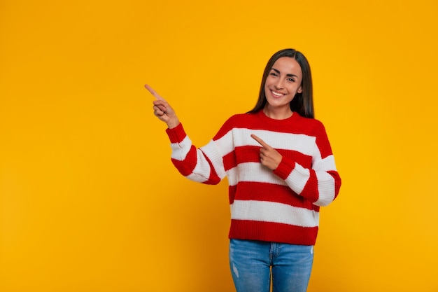 Mooie lachende jonge brunette vrouw in een trui wijst weg terwijl ze poseert geïsoleerd op gele achtergrond