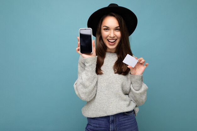 Mooie lachende jonge brunette vrouw met zwarte hoed en grijze trui geïsoleerd over blauwe achtergrond met creditcard en mobiele telefoon met lege display voor mockup camera kijken.