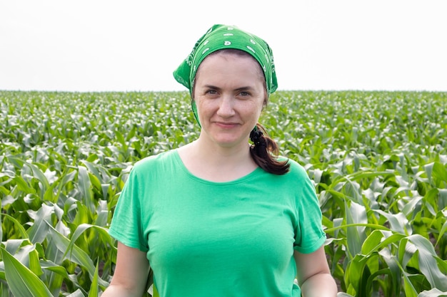 Mooie lachende jonge vrouw boer in groen t-shirt en groene zonnehoed staande in het midden van een groen maïsveld op een zonnige zomerdag buitenshuis. oogst tijd. biologische landbouw. zonlicht.