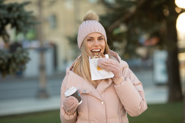 Mooie lachende jonge vrouw in warme kleding met een kop warme thee of koffie. Vrouw die koekjes eet openlucht.