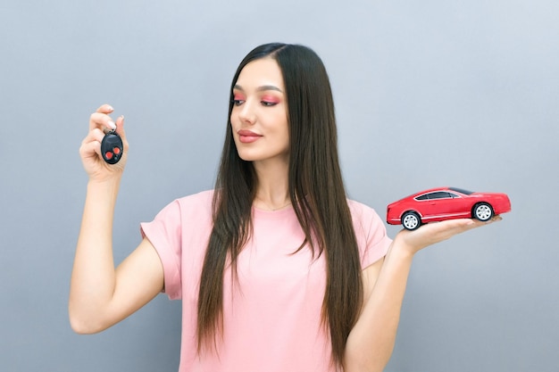 Mooie lachende jonge vrouw met autosleutels en een auto in haar hand het concept van leren in een rijschool en het kopen van een nieuwe auto op een grijze achtergrond