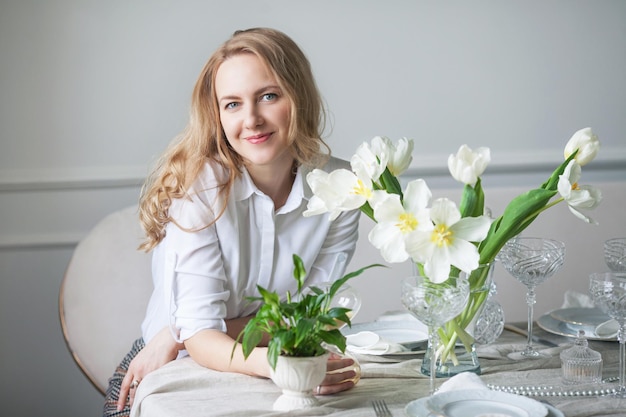 Mooie lachende jonge vrouw met lang blond haar zittend aan tafel in lichte witte kamer