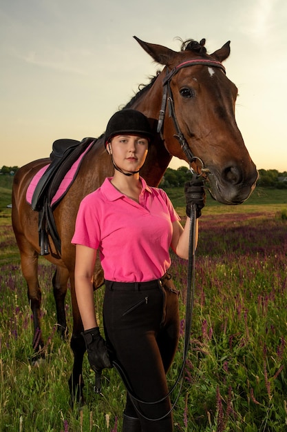 Mooie lachende meisjesjockey staat naast haar bruine paard met een speciaal uniform op een hemel en een groene veldachtergrond op een zonsondergang. Equitation sport competitie en activiteit.