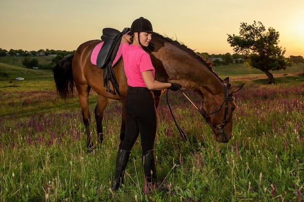 Mooie lachende meisjesjockey staat naast haar bruine paard met een speciaal uniform op een hemel en een groene veldachtergrond op een zonsondergang.