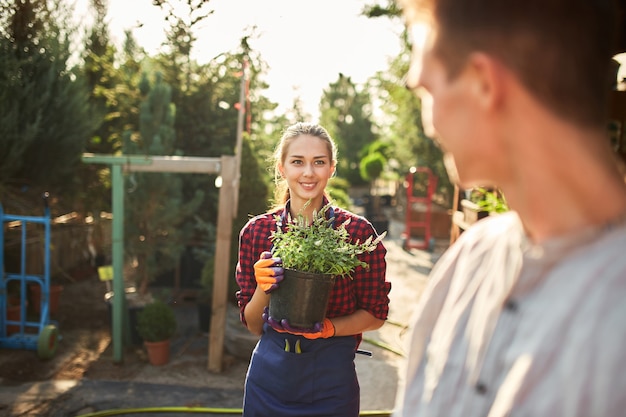 Mooie lachende meisjestuinman houdt pot met planten vast en kijkt op een zonnige dag naar de man in de prachtige tuin.