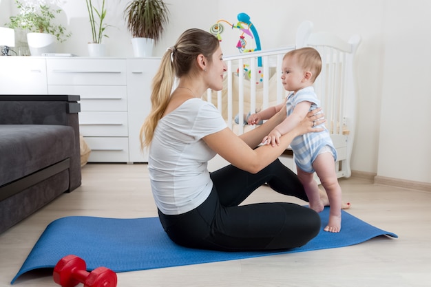 Mooie lachende moeder die yoga beoefent op fitnessmat in de woonkamer