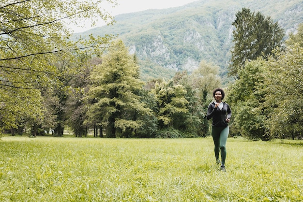Mooie lachende volwassen afro-amerikaanse vrouw loopt door groen gras in de buurt van het berglandschap.