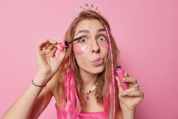 Mooie langharige Europese vrouw past mascara toe wil een mooi uitzicht hebben brengt hydrogel schoonheidspleisters onder de ogen aan om zakken te verwijderen draagt kroon en jurk geïsoleerd over roze achtergrond