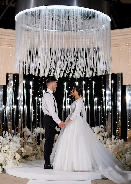 Mooie liefdevolle bruiden paar staande op luxe versierd met bloemen altaar 's nachts kijken naar elkaar elegante vrouw in lange gezwollen jurk met knappe man speciaal moment in het leven