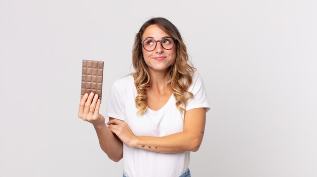 Mooie magere vrouw die haar schouders ophaalt, zich verward en onzeker voelt en een chocoladereep vasthoudt