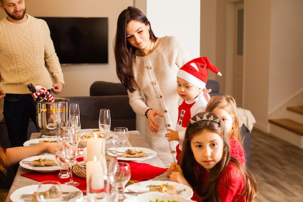 Mooie meisjes en schattige kleine jongen in kostuum van de kerstman zittend aan de kersttafel Kerst familiefeestje