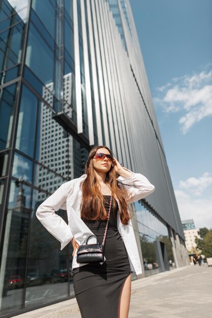 Mooie mode jongedame met zonnebril in een stijlvolle zwarte jurk met een wit overhemd met een handtas loopt door de stad in de buurt van een modern glazen gebouw