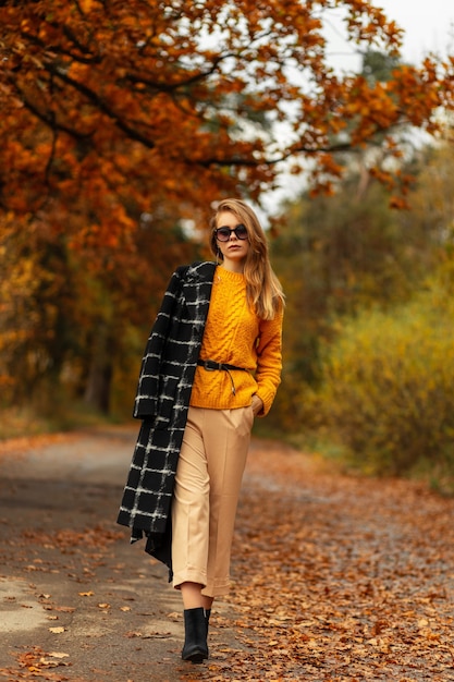 Mooie modieuze modelvrouw in trendy herfstkleding met een jas, gebreide trui, broek en laarzen loopt in het park met kleurrijke herfstbladeren