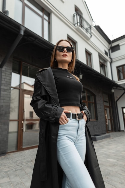 Mooie modieuze vrouw model met coole zwarte zonnebril in stijlvolle straatkleren met een trendy leren jas tanktop en hoog getailleerde jeans in de stad