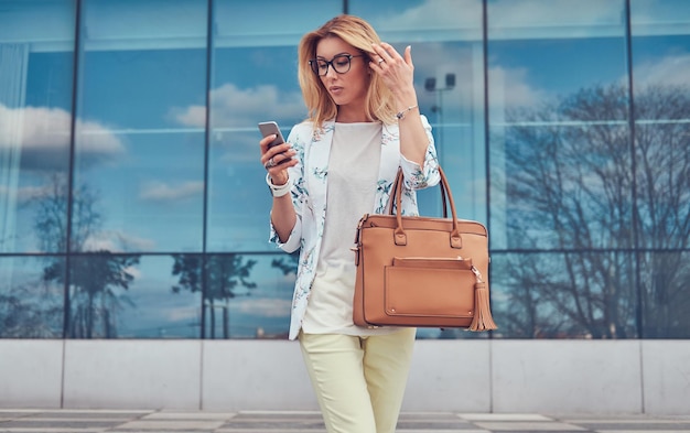 Mooie modieuze vrouwelijke blogger in stijlvolle kleding en bril met een handtas, met behulp van een smartphone, staande tegen een wolkenkrabber.