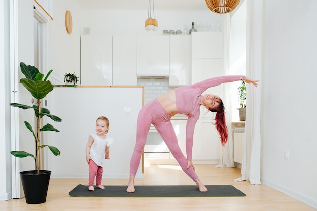 Mooie moeder doet rekoefeningen naast haar baby op een yogamat in de keuken