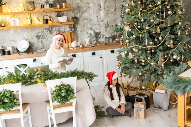Mooie moeder en dochter hebben plezier, knuffel en kus thuis bij de kerstboom in een wit interieur. Familiegeluk, vakantie, vreugde, vakantie, spelletjes met een vrouw. Nieuwjaarsvoorbereidingen