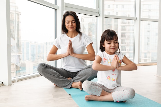 Mooie moeder en kind thuis mediteren, benen zittend op mat gekruist en yoga gebaar met palmen samen doen