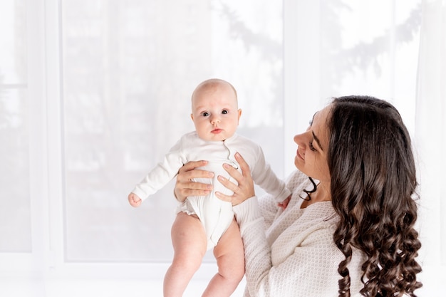 Mooie moeder met een baby in haar armen bij het raam van het huis, het concept van een liefdevolle en gelukkige familie, levensstijl