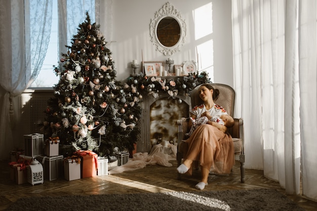 Mooie moeder zit in de fauteuil met haar kleine baby naast de open haard en de nieuwjaarsboom met geschenken in de lichte, gezellige kamer.