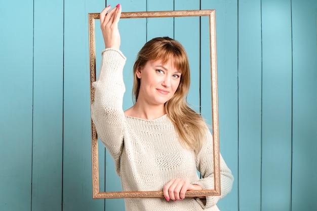 Mooie mooie blonde jonge vrouw in gebreide trui met losse haren houdt vintage houten afbeeldingsframe en glimlacht