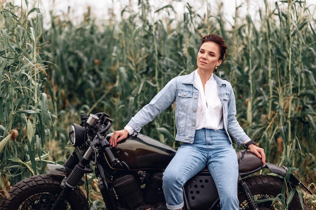 Mooie mooie jonge vrouw denim kleding dragen en zittend op een grote zwarte retro motorfiets tegen een achtergrond van gras en groen. Concert van snelheid