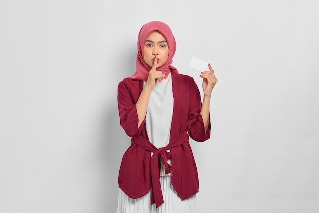 Mooie moslimvrouw in casual shirt en hijab met creditcard, vinger die lippen bedekt, maakt stiltegebaar geïsoleerd op witte achtergrond. mensen religieuze levensstijl concept