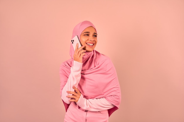 Foto mooie moslimvrouw in roze hijab praten aan de telefoon en glimlachend op een beige achtergrond