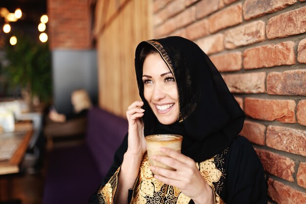 Mooie moslimvrouw in traditionele slijtage die in cafetaria zit en van koffie geniet.