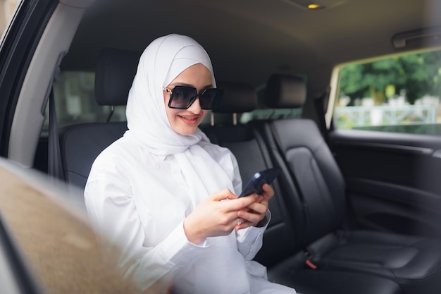 Mooie moslimvrouw met een witte hijab die op de achterbank van een auto zit en smartphone gebruikt