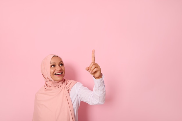 Mooie moslimvrouw van Midden-Oosterse etniciteit gekleed in religieuze outfit en bedekt hoofd met hijab etniciteit glimlacht en kijkt op en wijst haar wijsvinger omhoog op een kopie ruimte van roze achtergrond