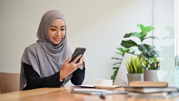 Mooie moslimzakenvrouw die hijab draagt, zit aan haar werktafel en gebruikt een smartphone