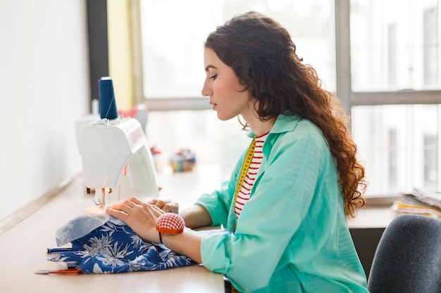 Mooie naaister in kleurrijk shirt dromerig met behulp van naaimachine met groot raam op de achtergrond in moderne naaiatelier