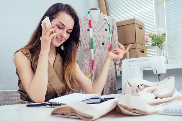 Mooie naaister met lang haar schrijft in een notitieboekje. de kleermaker maakt een verzameling outfits en praat telefonisch met de klant. jonge vrouw merkkleding notities ideeën in notebook