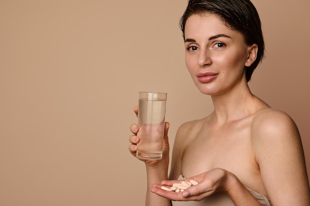 Mooie natuurlijke schoonheidsvrouw met een gezonde gloeiende heldere huid die naar de camera kijkt met een medicijn, een complex van vitamines en mineralen in farmaceutische dragees en een glas water in haar handen