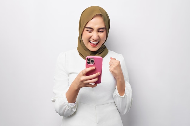 Mooie opgewonden aziatische moslimvrouw die hijab draagt met behulp van mobiele telefoon typend sms-bericht doet winnaargebaar geïsoleerd op witte achtergrond studioportret mensen religieus levensstijlconcept