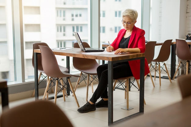 mooie oude vrouw in een rode jas werkt op kantoor met een laptop