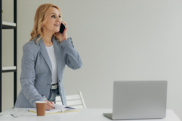 Mooie oudere zakenvrouw die op slimme telefoon spreekt terwijl ze werkt en notities schrijft in Kladblok Rijpe vrouw 50s