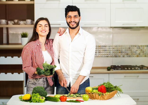 Mooie paar plezier als koken knappe bebaarde man komkommer snijden mooie donkerharige vrouw met broccoli in de hand concept van gelukkig gezinsleven en gezonde voeding