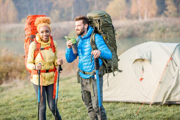 Mooie paar wandelen met kleurrijke rugzakken op het groene gazon met tent op de achtergrond, man drinkwater uit de fles