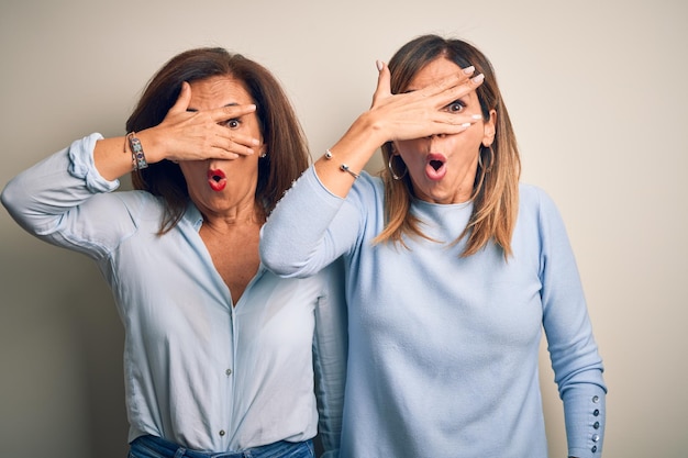 Foto mooie paar zussen van middelbare leeftijd die over een geïsoleerde witte achtergrond staan en geschokt gluren over gezicht en ogen met de hand kijkend door vingers met een beschaamde uitdrukking
