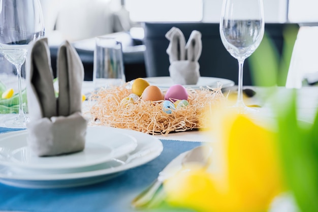 Mooie paastafel met feestelijk decor binnenshuis Servet in de vorm van konijnenoren kleurrijke eieren in nest verse gele tulpen in vaas Zonlicht ansichtkaart groet Interieur