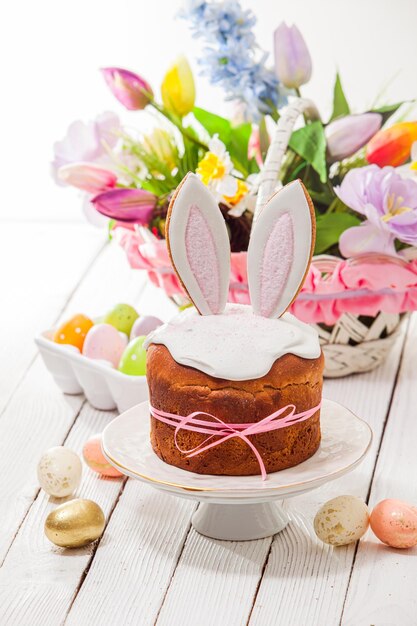 Mooie Pasen taarten bedekt met witte suiker slagroom en versierd met konijnenoren geplaatst op witte taart staan Mand met bos bloemen op achtergrond kleurrijke eieren wit houten oppervlak