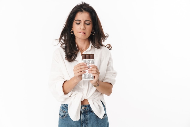 Mooie peinzende jonge brunette vrouw die vrijetijdskleding draagt, geïsoleerd over een witte muur staat en chocoladebord vasthoudt