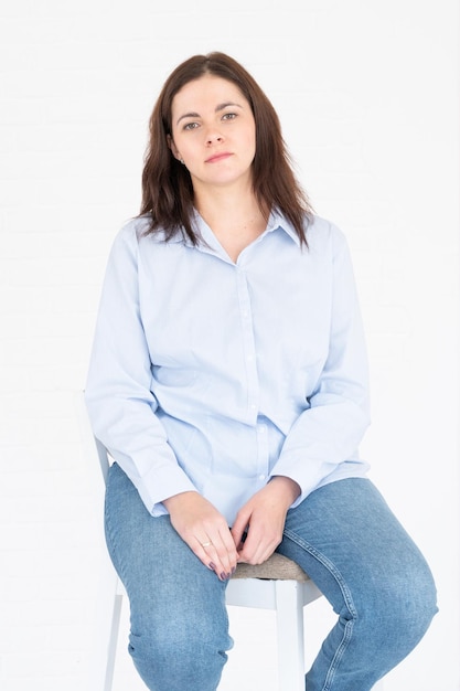 Mooie plus size model vrouw in shirt en spijkerbroek poseren in studio op stoel, witte achtergrond met kopie ruimte.