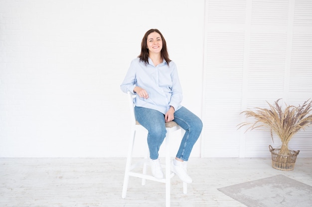 Mooie plus size model vrouw in shirt en spijkerbroek poseren in studio op stoel, witte achtergrond met kopie ruimte.