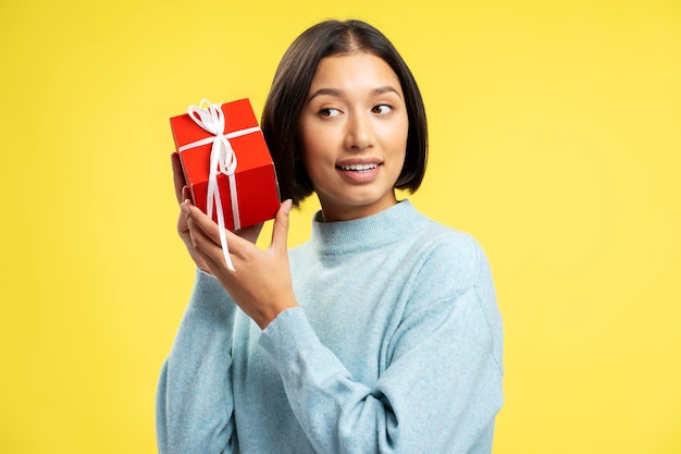 Foto mooie positieve aziatische vrouw met een rode geschenkdoos met lint die wegkijkt en geïsoleerd staat op een gele achtergrond concept van verjaardagviering