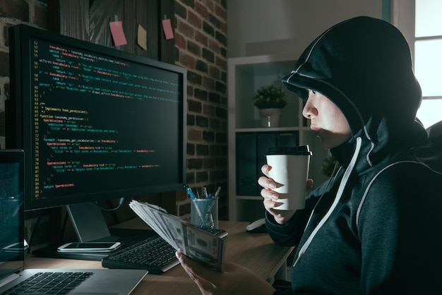 mooie programmeur-hacker die hete koffie drinkt, ontspannen en contant bankbiljet vasthoudt, kijkend naar computerscherm en toezicht houden op online virusverspreiding.