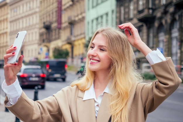 Mooie rijke casual blonde stijlvolle mode zakenvrouw met een telefoon in haar hand maken selfie in Europese gebouwen en straat.