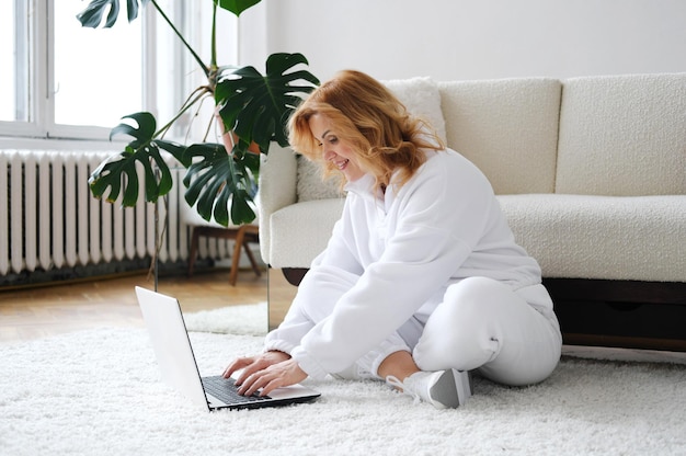 Mooie rijpe blonde vrouw zit in de woonkamer op het tapijt en gebruikt een laptop Het concept van onderwijs na 40 jaar opleidingen en een nieuw beroep